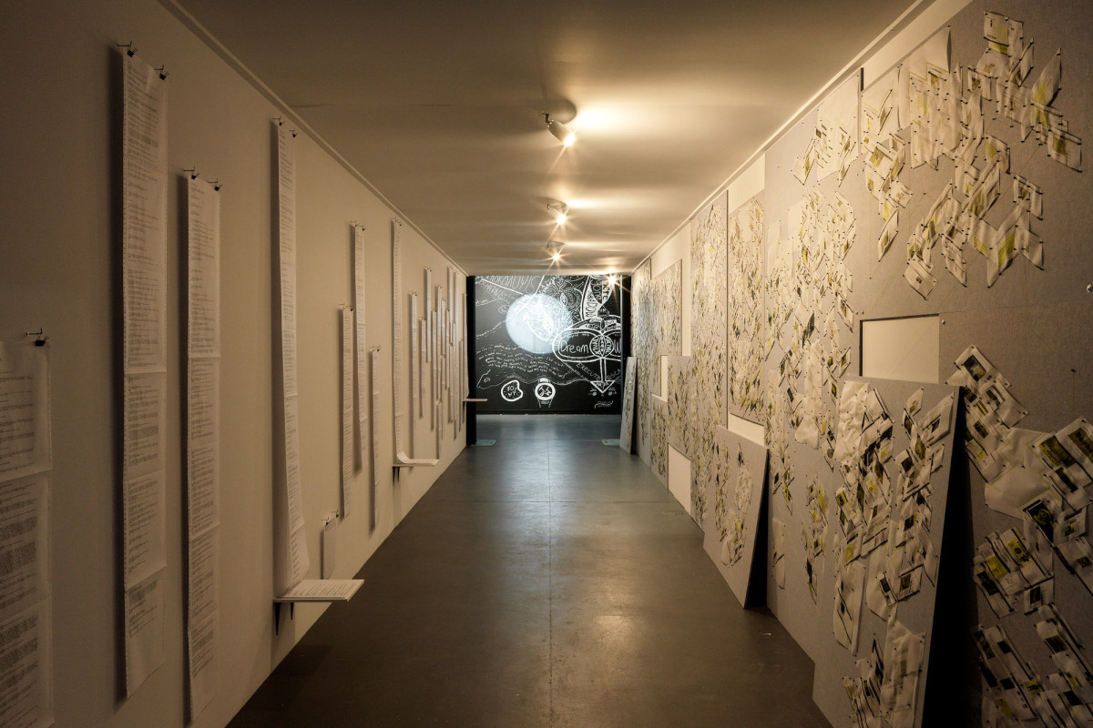 A tunnel is lined with printer paper to the left and images diagrams to the right. The wall is painted black with white writing at the end of the tunnel.
