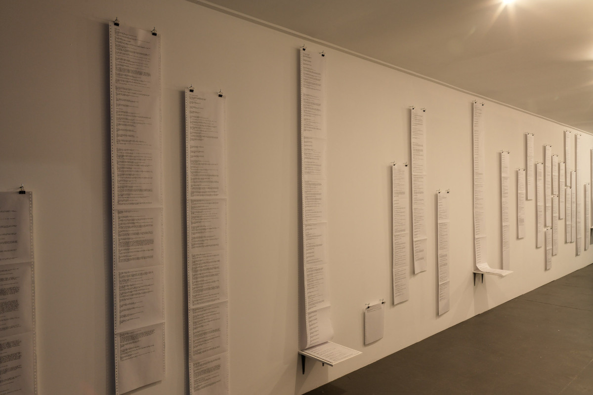 Printer paper with writing vertically lines the gallery wall.