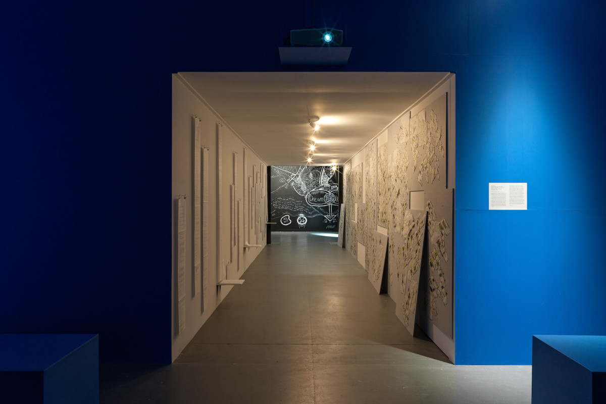 A view of a brightly lit tunnel with pages of writing and diagrams by Roy Ananda. At the end of the tunnel Gilselle Stanborough's painted installation.
