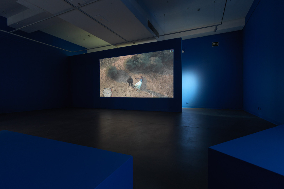 Film still of two people in the bush projected onto the blue, dimly lit gallery space.