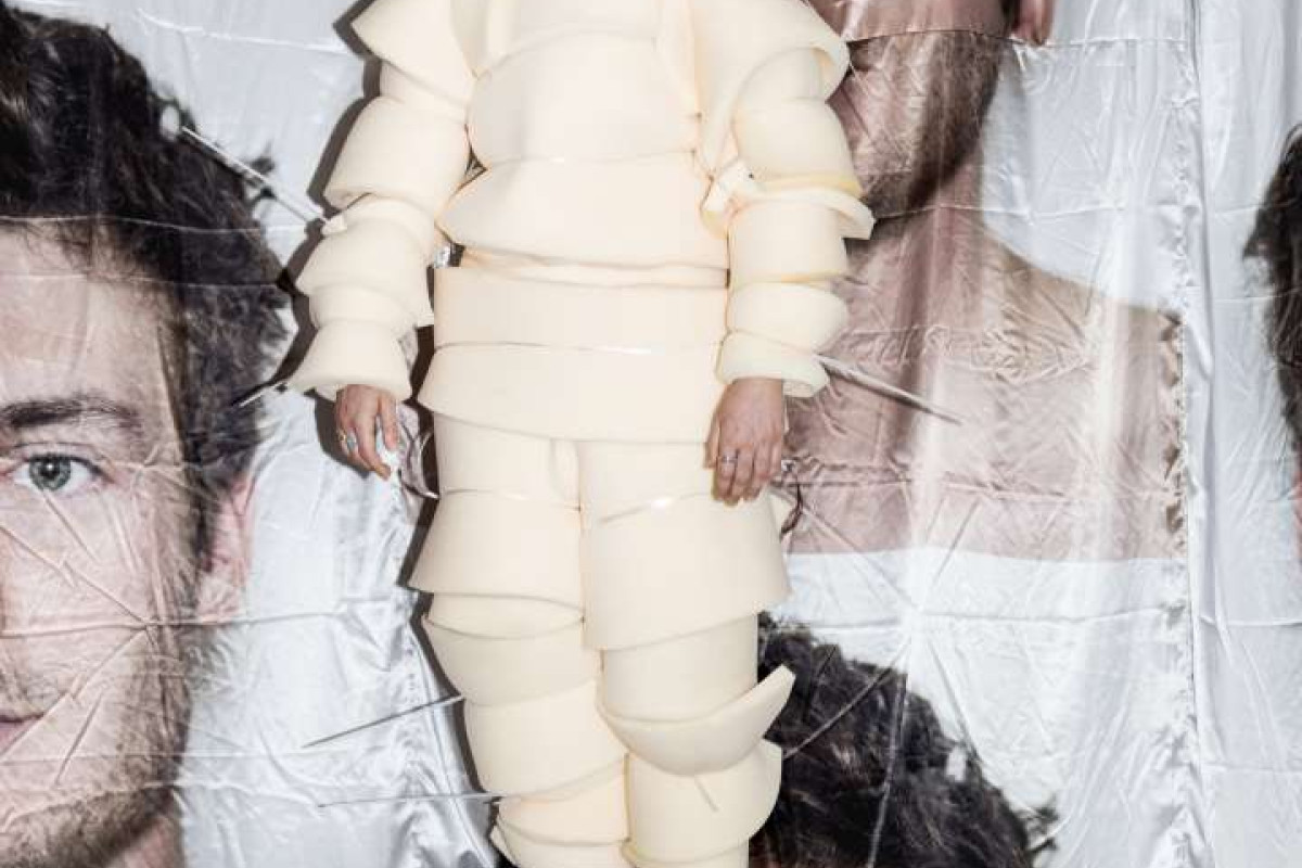 A person, wearing a suit made of foam pieces, stands in front of a draping fabric with a mans face printed on it.