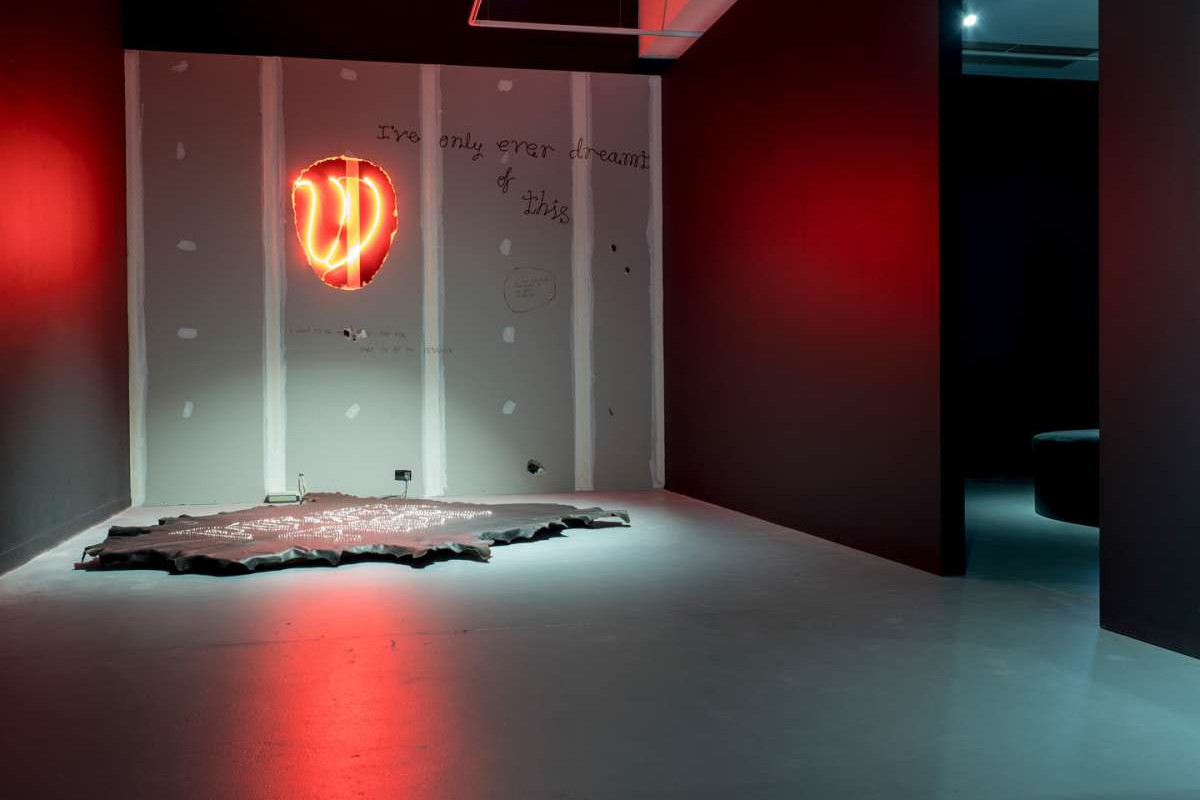 A dark room is lit only by a redneon light just visible through a hole in plaster walls, on the wall hand-written is the text 