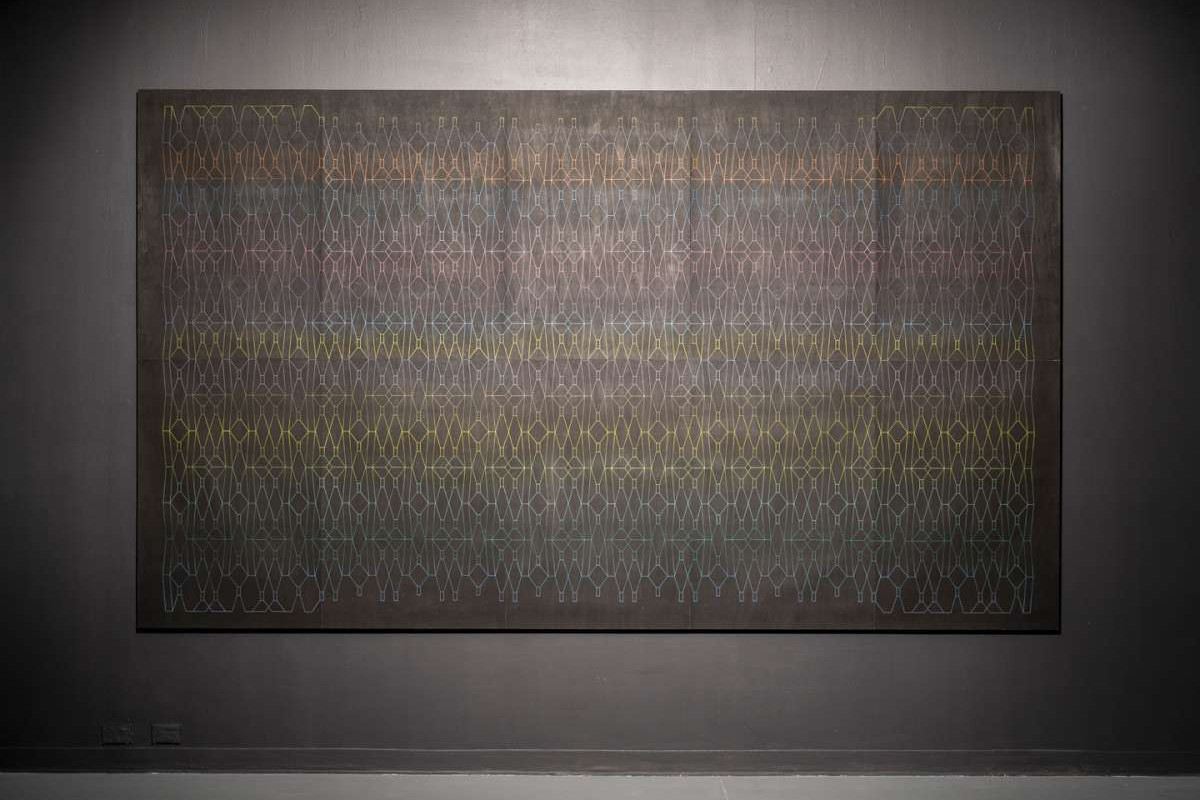 A large great canvas with multicoloured geometric line drawings done in oil pastel, hangs on a grey wall.