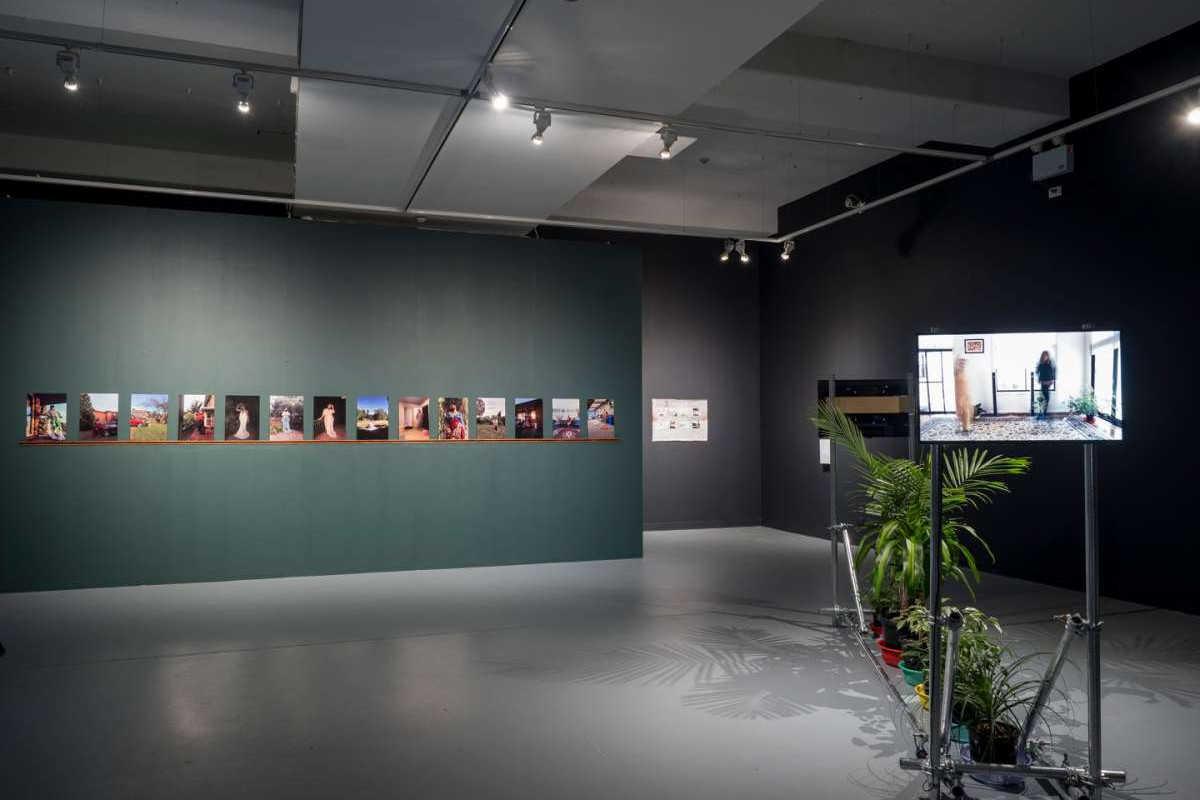 A wide shot of the ACE main gallery shows a dark green wall with 14 photographs on it, and a tv screen in the foreground, with a row of house plants lined up behind it.