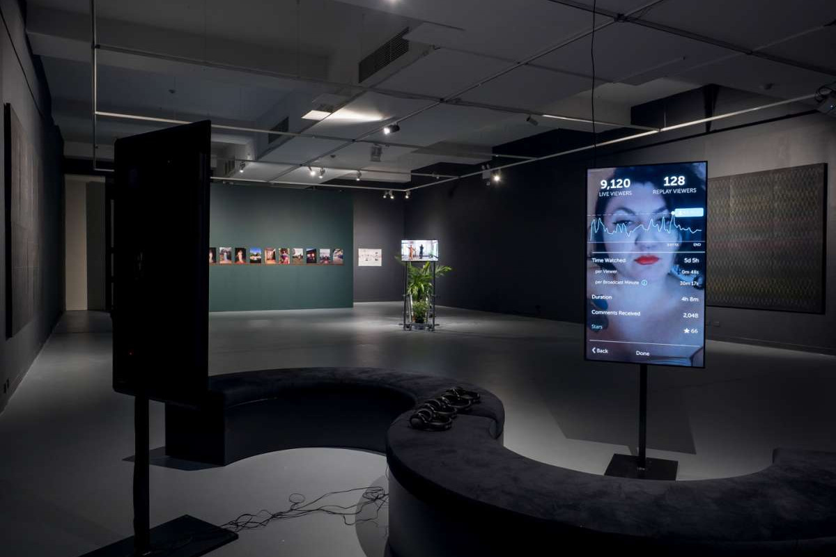 A widest of the ACE main gallery shows a curved seat in front of a tv screen with a woman's face and statistics and graphs over top of it, in the background a green wall with artwork hanging on it is visible.