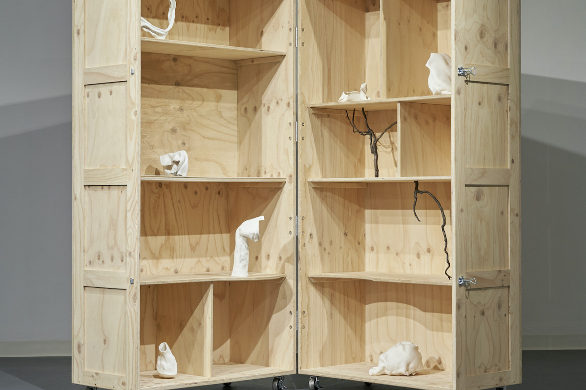 A large open book shaped wooden cupboard on wheels. Abstract white objects and wooden sticks sit on shelves.