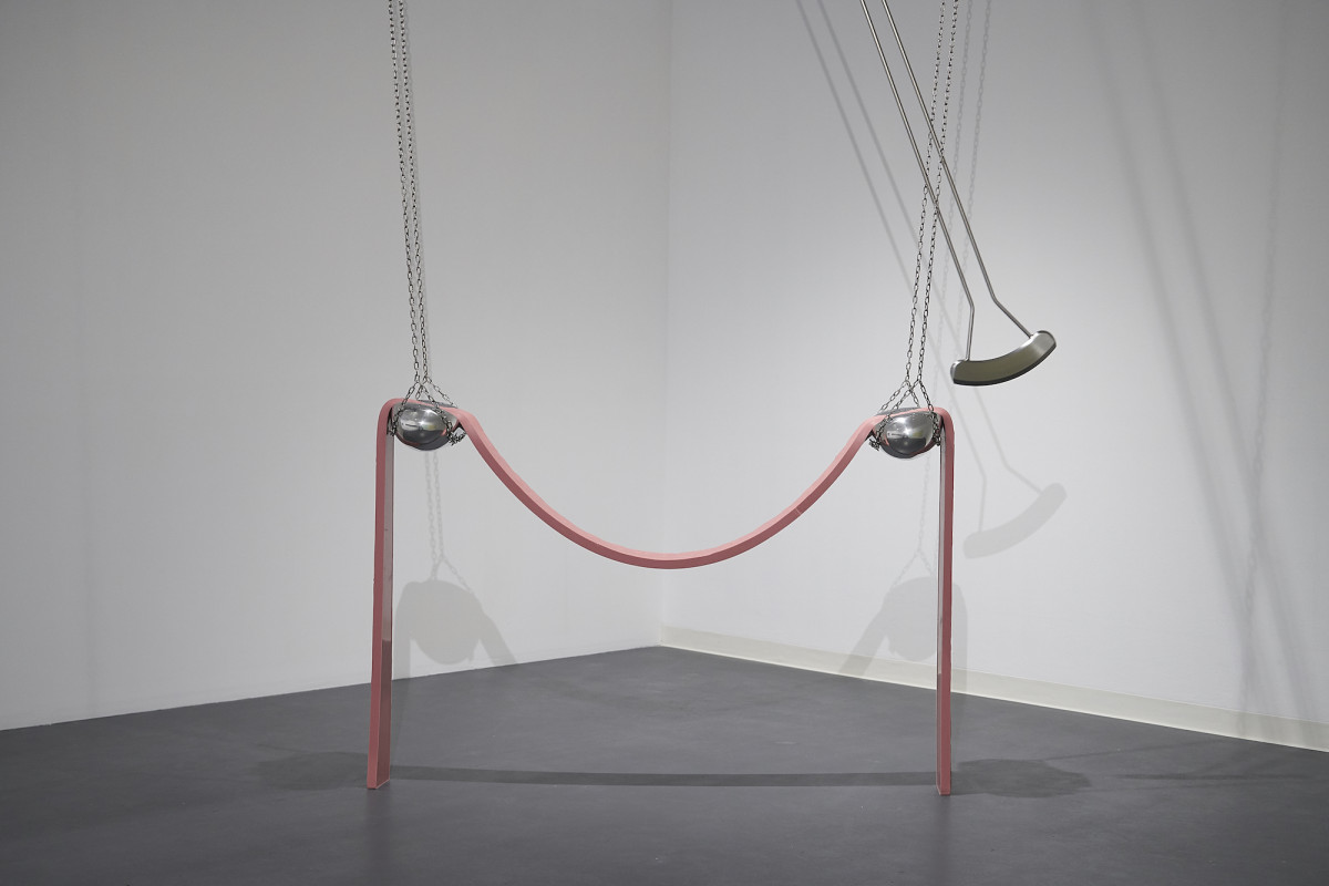 Sculpture consisting of two metal, chained, hanging oblong objects with a pink rubber strip draped over. A swinging metal pendulum swings closely back and forth close to the oblong objects.