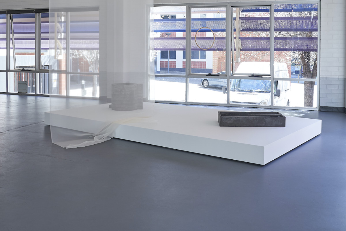 Brightly lit gallery large white plinth with rectangular and circular metal sculptures on top. Hanging yellow sculpture seen behind white, fabric, curtain veil from celling to floor.