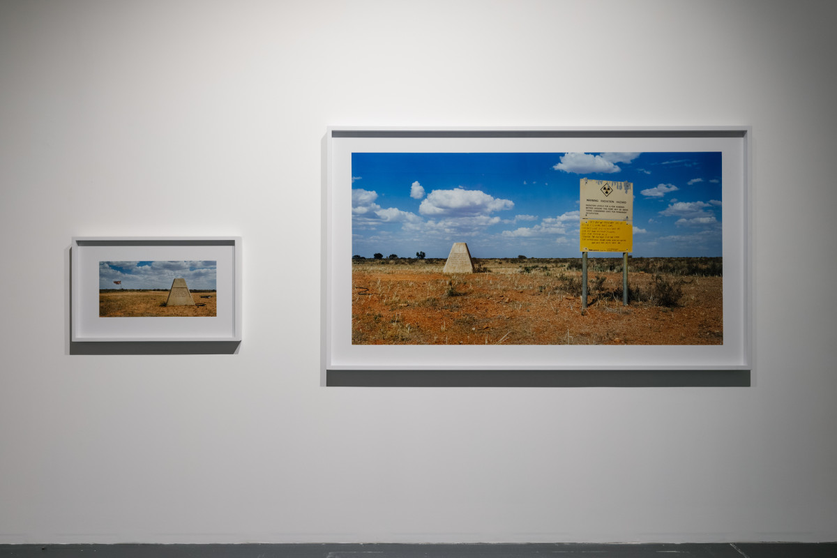 Two photographs framed side by side the right is a larger image with a stone monument and yellow sign in red dessert with blue skies. The smaller, framed image to the left is a close up of the stone monument.