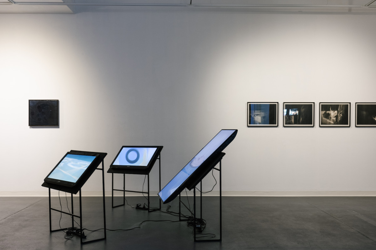 Image of gallery with three tv screens in a circular pattern. There are paintings hung landscape 1m up the wall in the background.