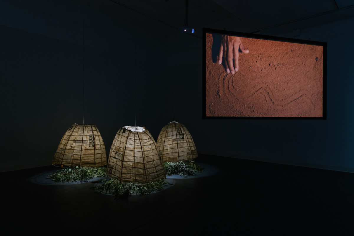 Dark gallery with three internally Illuminated lamp, shelters with gum leaves below. A large screen to the right displays a hand drawing squiggles in red sand.