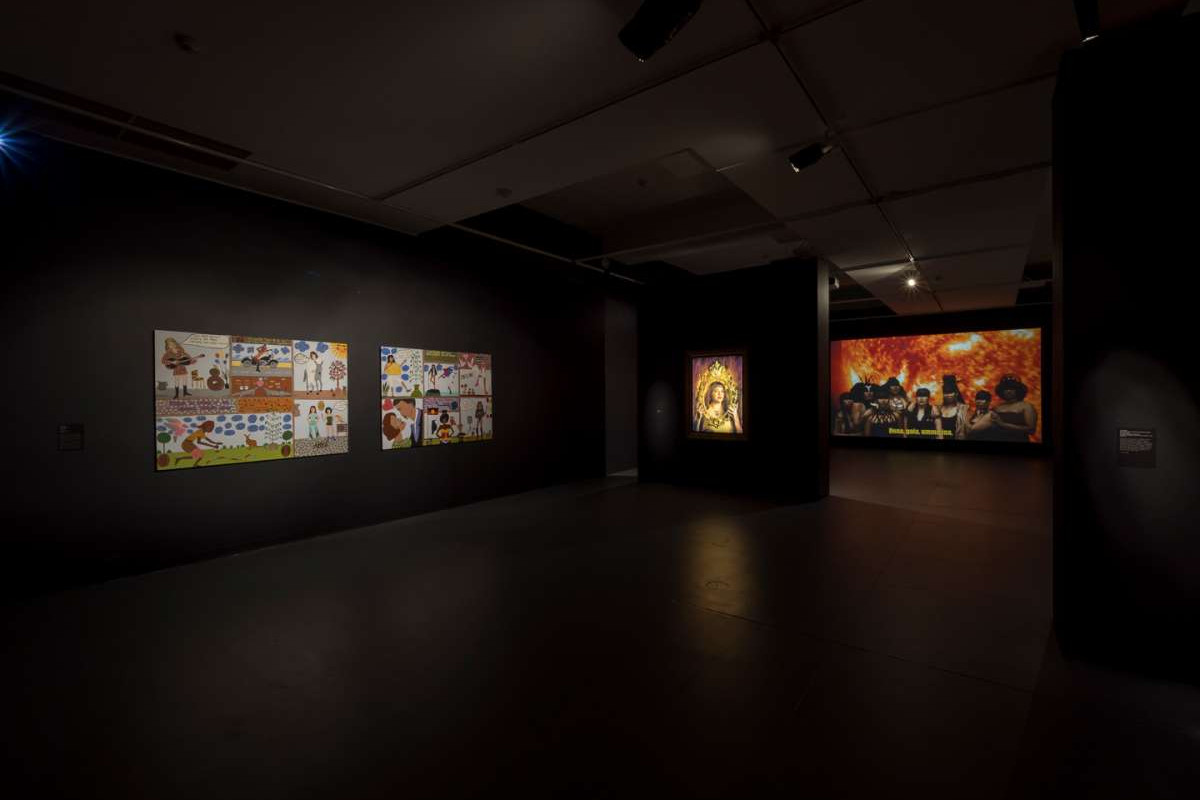 A wide shot of the ACE main gallery, with paintings, a photograph and projections on all of the black walls.