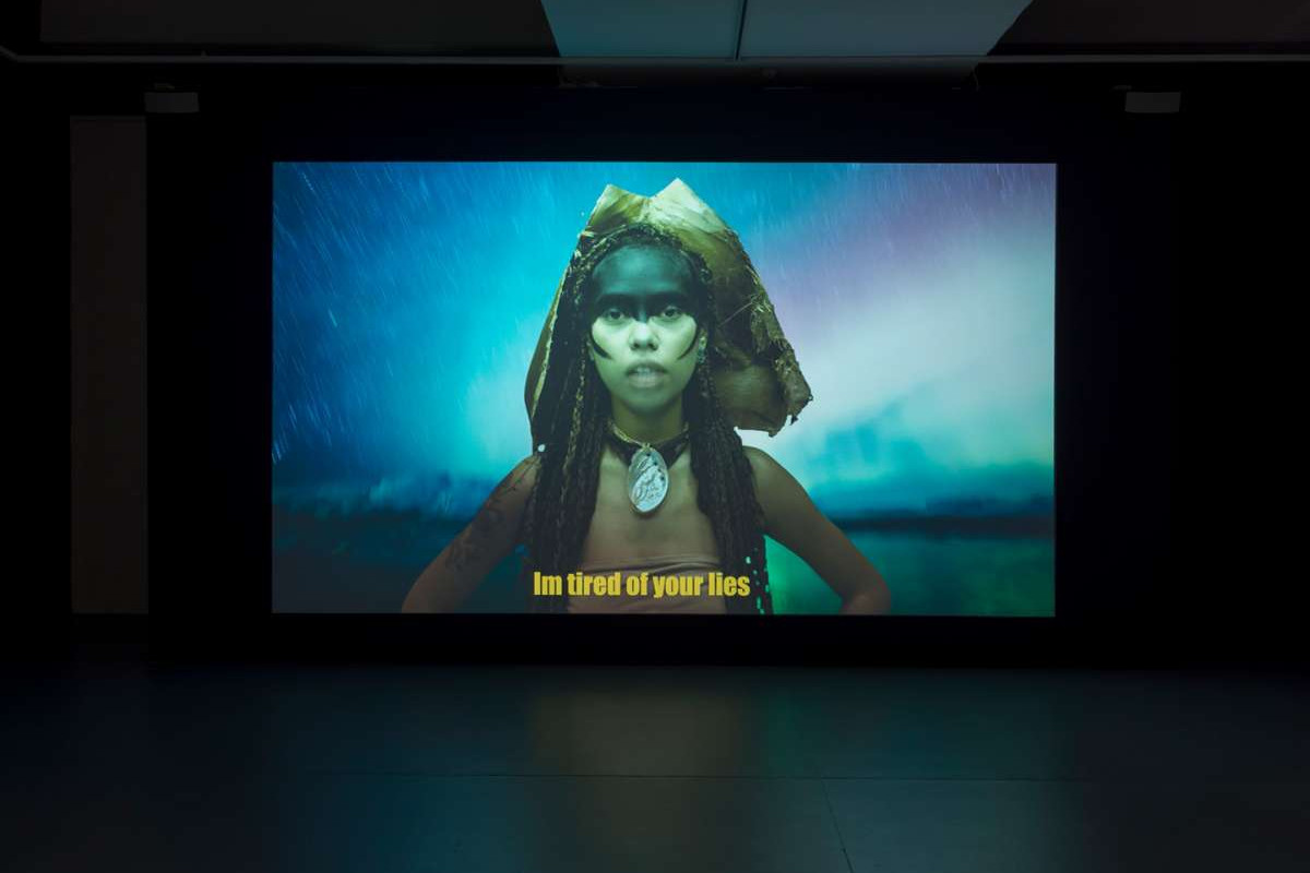 A Video still of a woman with her face painted, wearing a headpiece and a large necklace, standing in front of a dreamily coloured night sky.