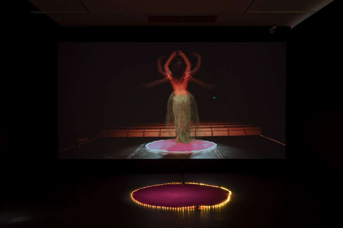 A video still of a woman, blurred in motion, while dancing to an empty theatre in a grass skirt.