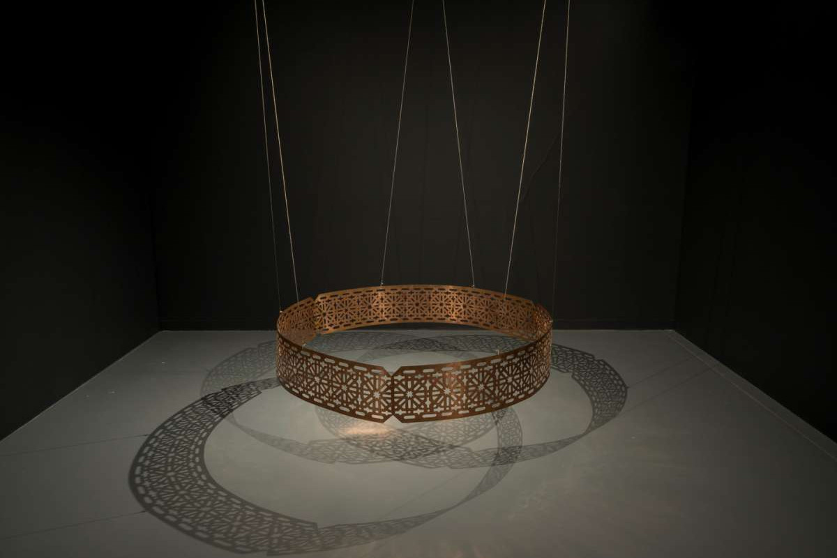 A circular copper sculpture, with detailed geometric cutout patterns on it, is suspended just above the ground