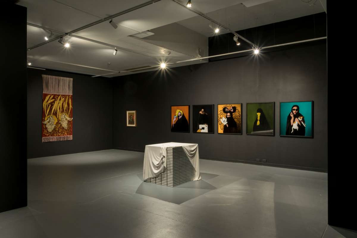A black wall with seven seperate pieces of art hanging on it, in the centre of the room sits a white cube with fabric draped over it.