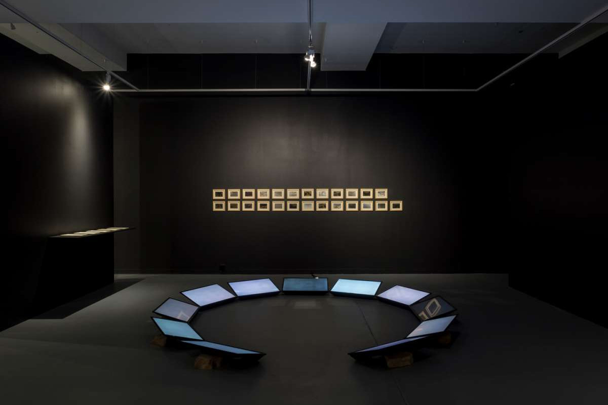 11 lit up screens sit in a circle in the centre of a black room, behind them on the wall is a series of 25 small gold paintings.