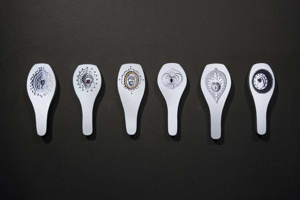 Six white sculpted artworks with detailed, layered hand cut out patterns, sit side by side on a black wall.