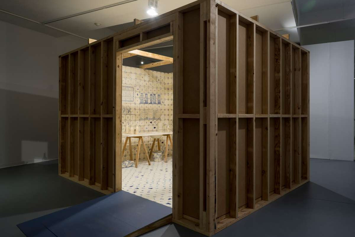 A small room made of wood with an open door, you can see inside the room is tiled from floor to ceiling with ceramic white tiles with blu drawings on them.