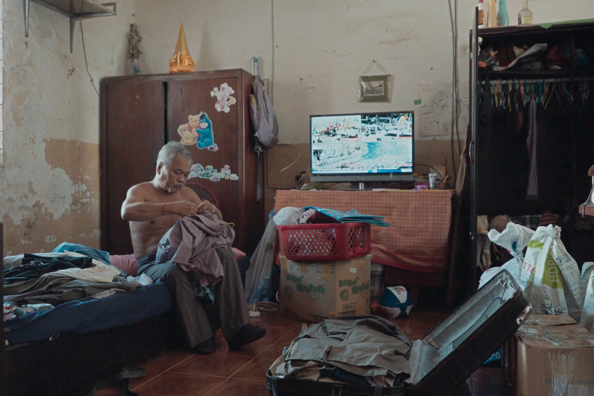 Man sits on bed in bedroom, buttoning shirt with a suitcase open in front of him.