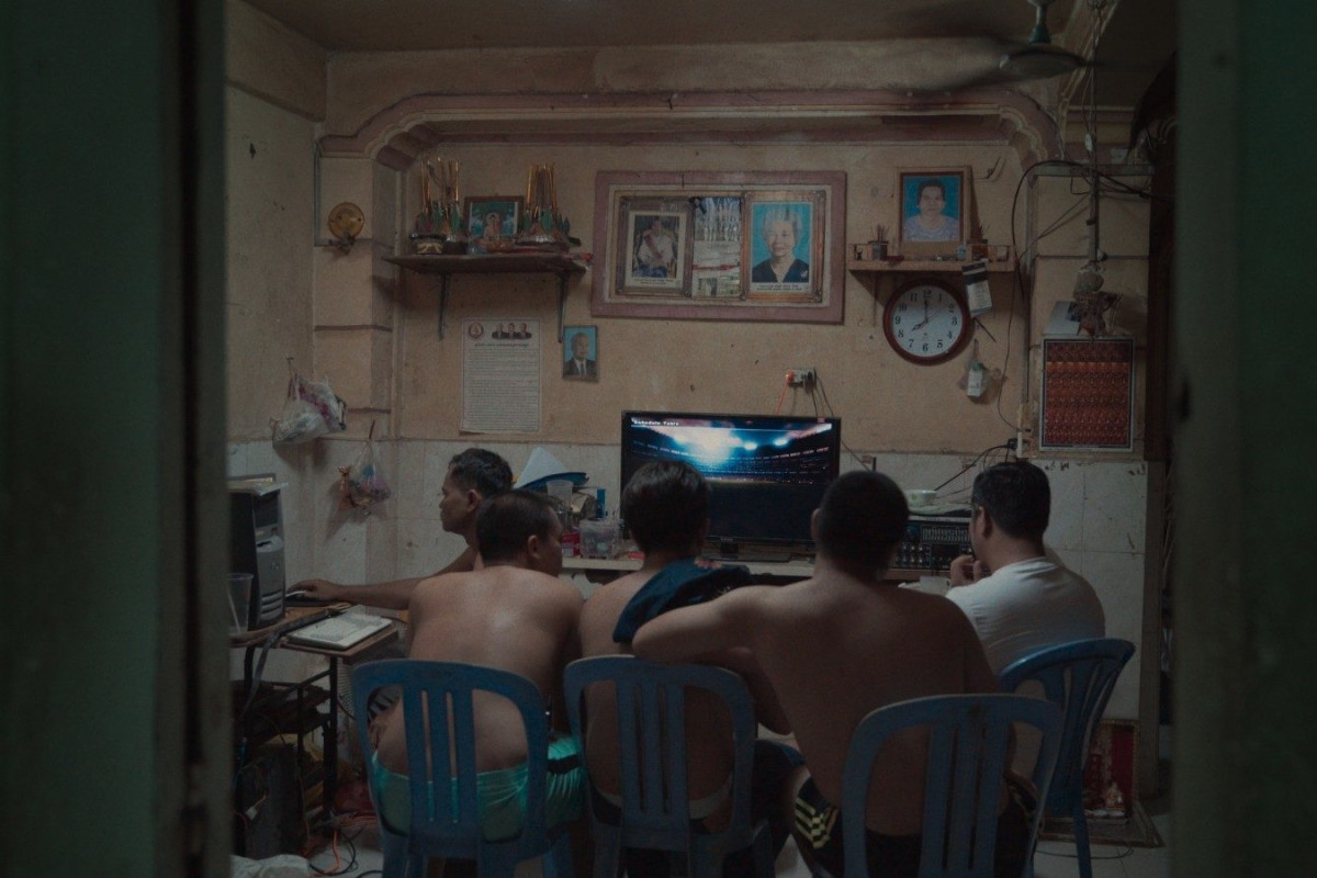 Four men watch a tv screen in a dim room, while another man is on a computer.
