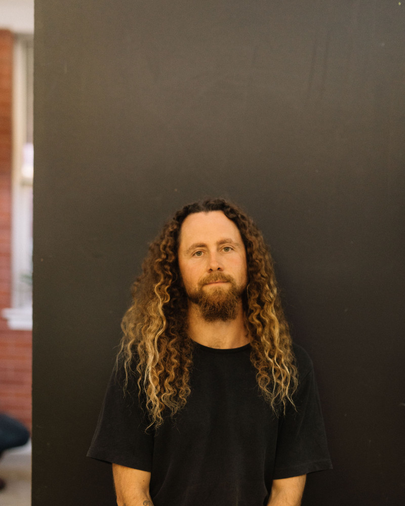 Artist Brad Darkson sits in a front of a black wall. He has long curly light brown hair, a beard and wears a black t-shirt.