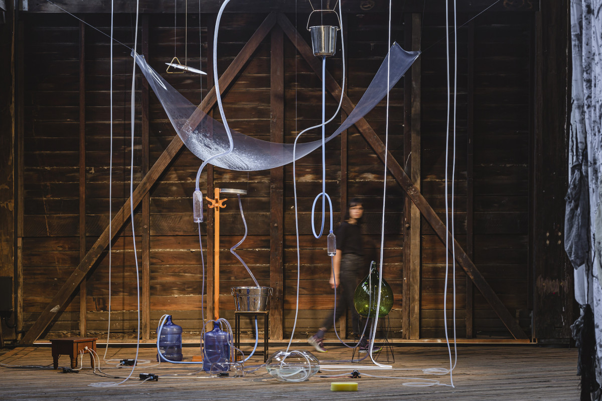 Various objects are joined by plastic pipes containing water, to create one large suspended installation. A person walks behind the installation.