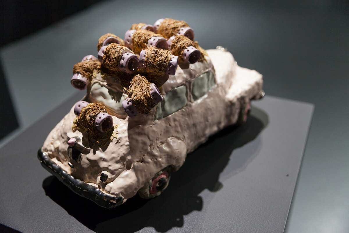 A hand sculpted model car with hair curlers on top of it sits atop a black plinth.