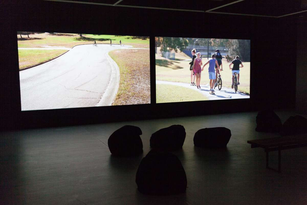 Two video projections sit side by side, on the screens are young people riding bikes.