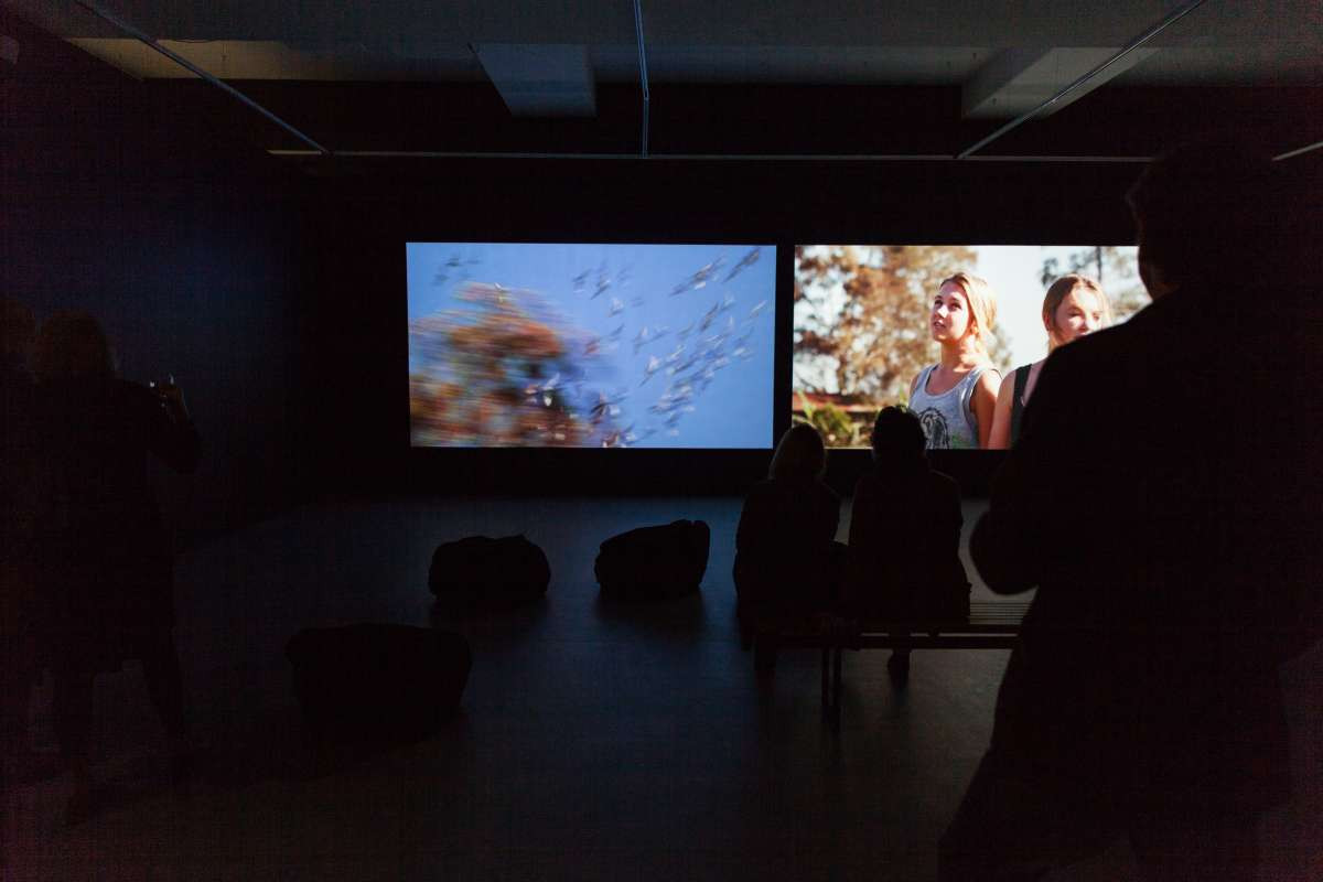 Two video projection sit side by side, on the screens are young girls. In the foreground are silhouettes of people watching the screens,