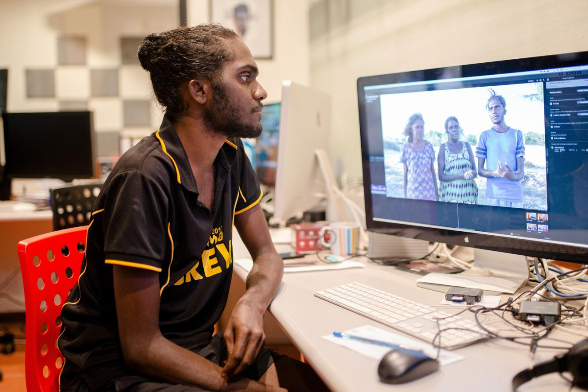 Man sits behind desk, looking at computer screen displaying photograph of three people.