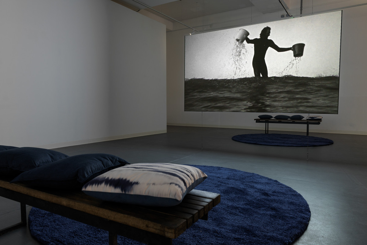 Suspended projection screen in ACE gallery, displaying image of surfer in ocean holding a bucket in each hand.