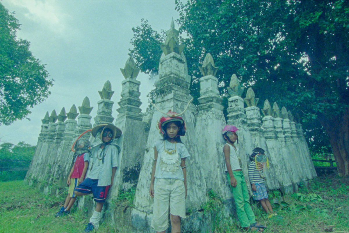 A group of 5 children stand in front of a row of tall, weathered stone spires in an outdoor setting. The children wear colourful hats and casual clothing, with trees and grass surrounding the scene.