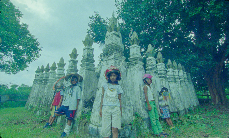 A group of 5 children stand in front of a row of tall, weathered stone spires in an outdoor setting. The children wear colourful hats and casual clothing, with trees and grass surrounding the scene.