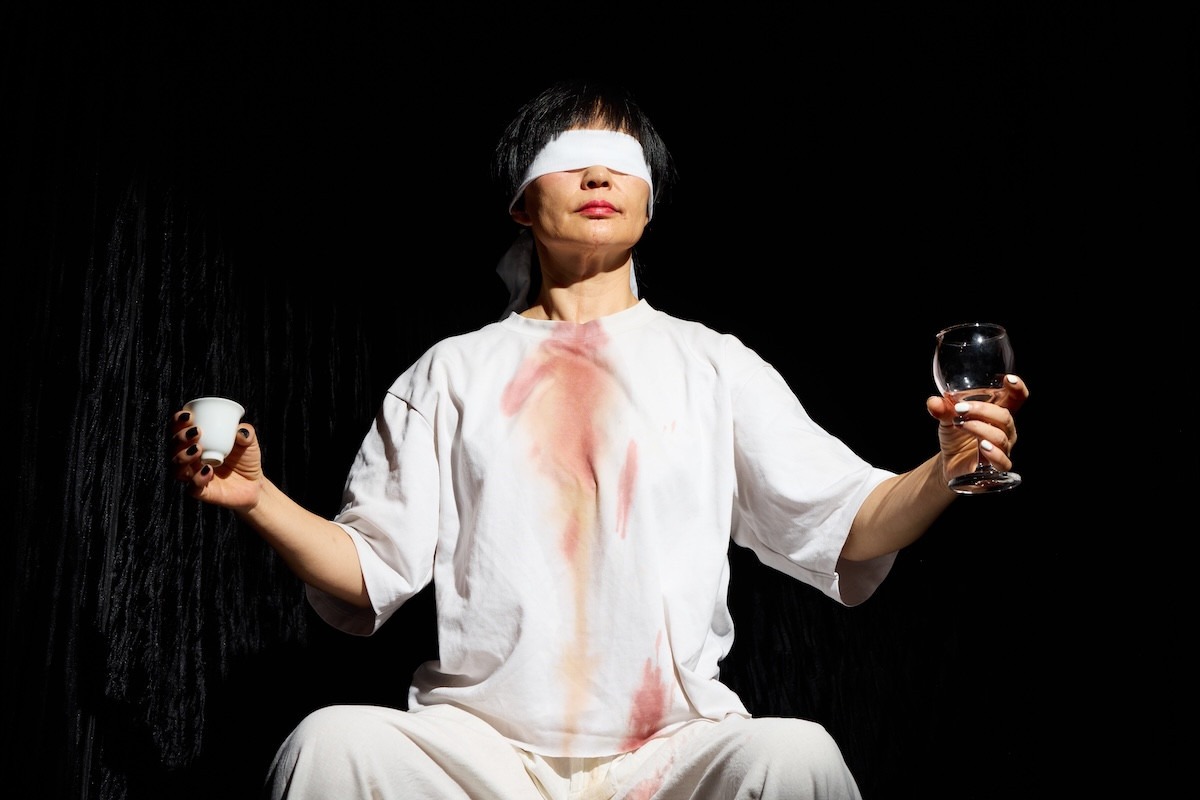 Spotlight on blindfolded person holding out cup of tea in right hand and wine glass in left hand on a dark stage.