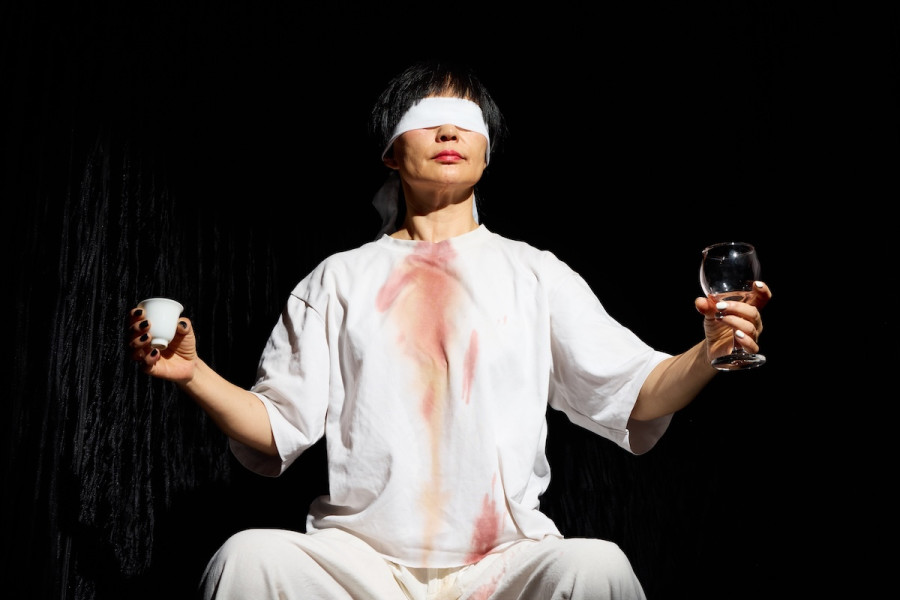 Spotlight on blindfolded person holding out cup of tea in right hand and wine glass in left hand on a dark stage.