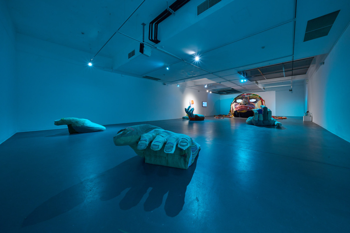 A wide photograph of the gallery space, in which inhabits Rockamora's head, feet and hands. The space is lit with blue light.