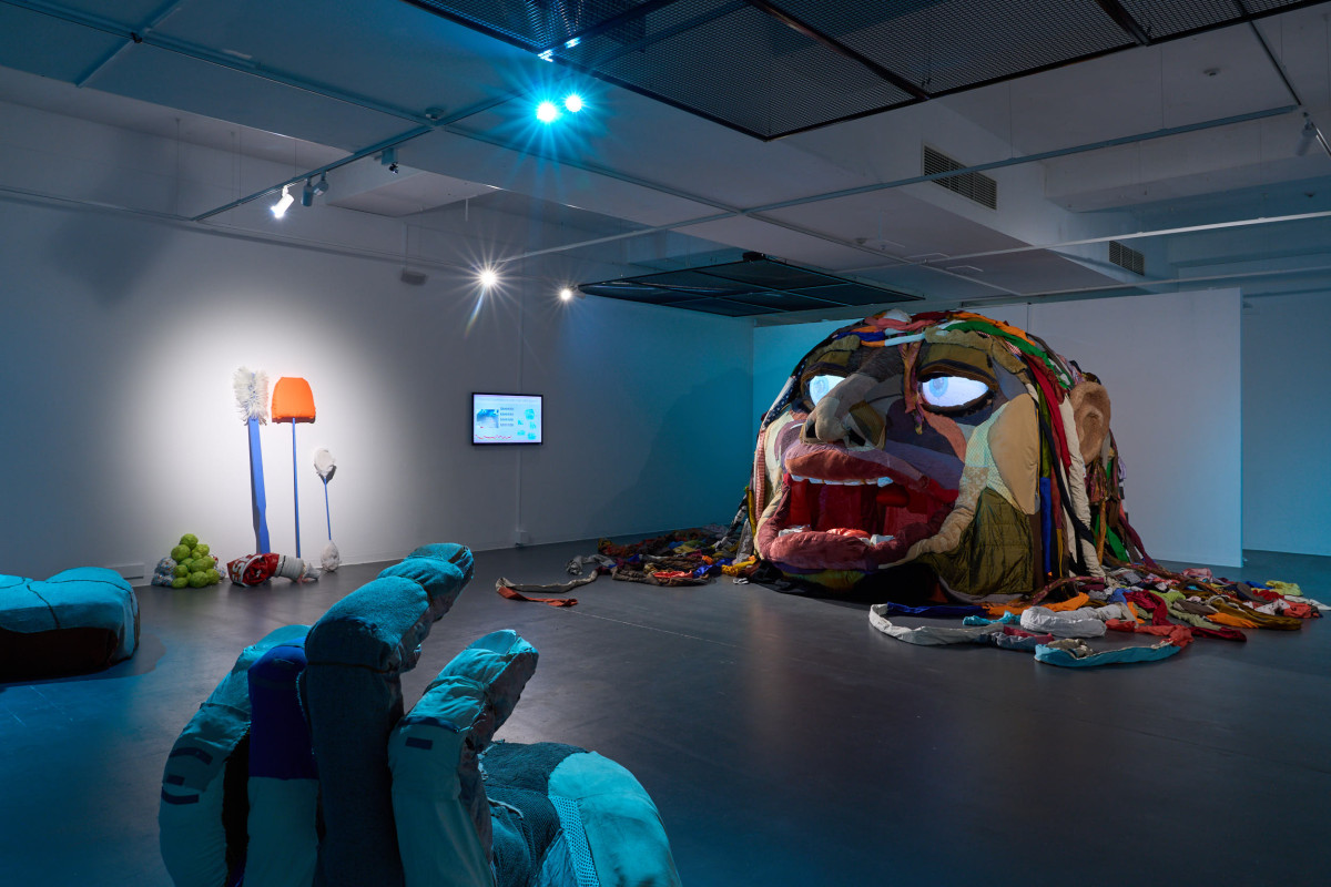 A photograph of the gallery space inhabited Rockamora's head, hands, a screen, and large tools. The space is lit with blue light.