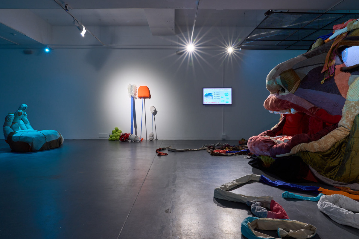 A photograph of the gallery space inhabited Rockamora's head, a hand, a screen, and large tools. The space is lit with blue light.