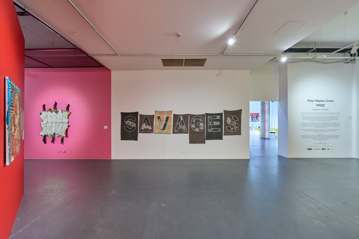 A wide view of the PRIDE exhibition in ACE's gallery. Focus is on a work that features the word 'SAVAGES' across 7 tapestries hanging on the North gallery wall.