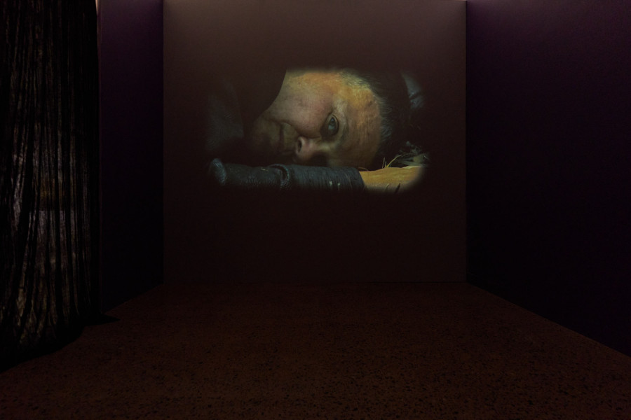 Video projection in a dark room showing a close-up of a person lying down, gazing toward the camera.