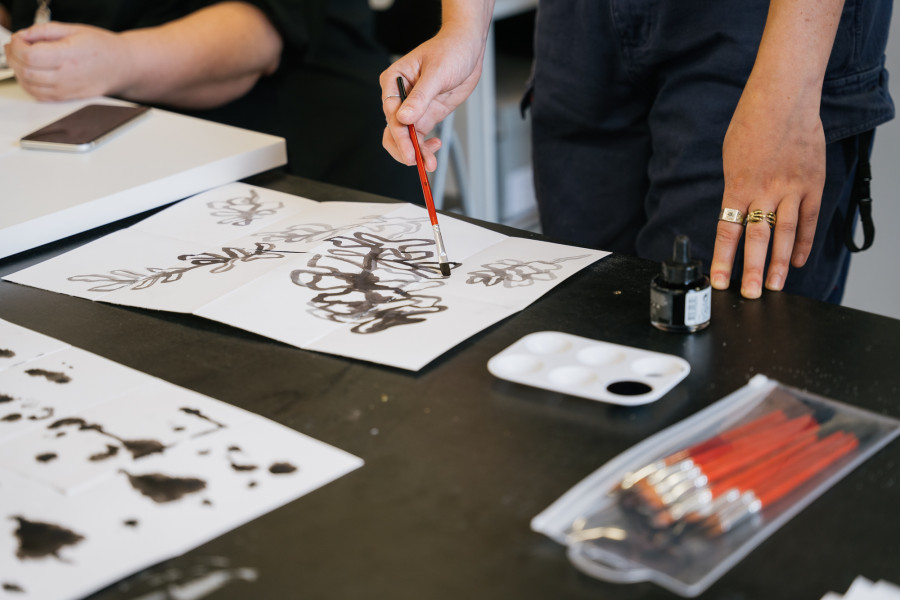 A close-up of Asha's hands painting during their workshop. Asha uses a thin paintbrush and paints black ink onto white paper.