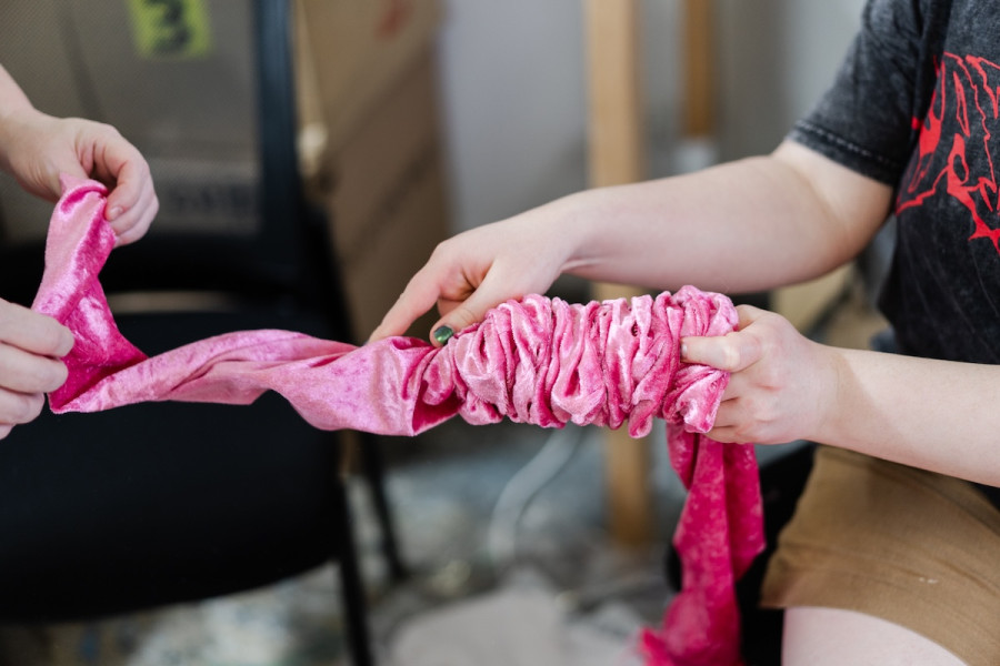 Two people manipulating a piece of bright pink, ruched fabric. One person tightly gathers the material while the other holds the end. The scene takes place in a workshop setting.