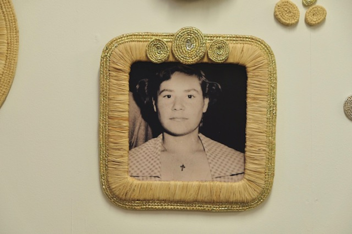 Artwork by Carly Tarkari Dodd 'Nina Dodd'. A square old photograph of a woman is framed. The frame made from raffia and ribbon. 6 smaller old black & white photographs are spread across the white surface behind the frame. One photograph sits on top of the frame on the left hand side.
