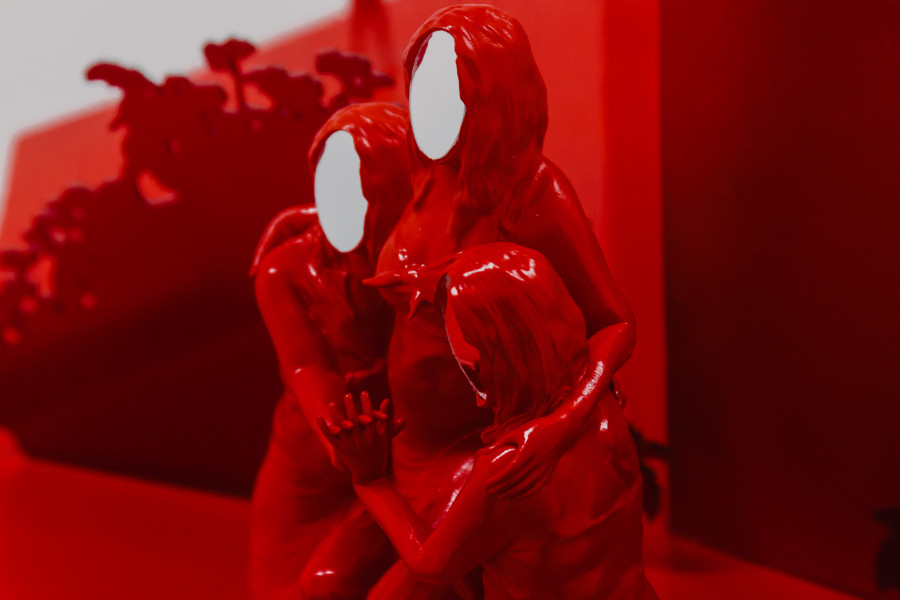 A bright red sculpture of three woman embracing, their faces made of mirror
