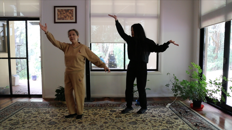 The artist and her mother stand in a lounge room flanked by pot plants, holding their arms in an identical outstretched pose. Video still by Eugene Choi.