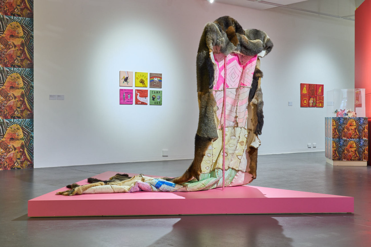 A large cloak made from possum pelts with a rainbow inside, stands on a pink plinth in a gallery.