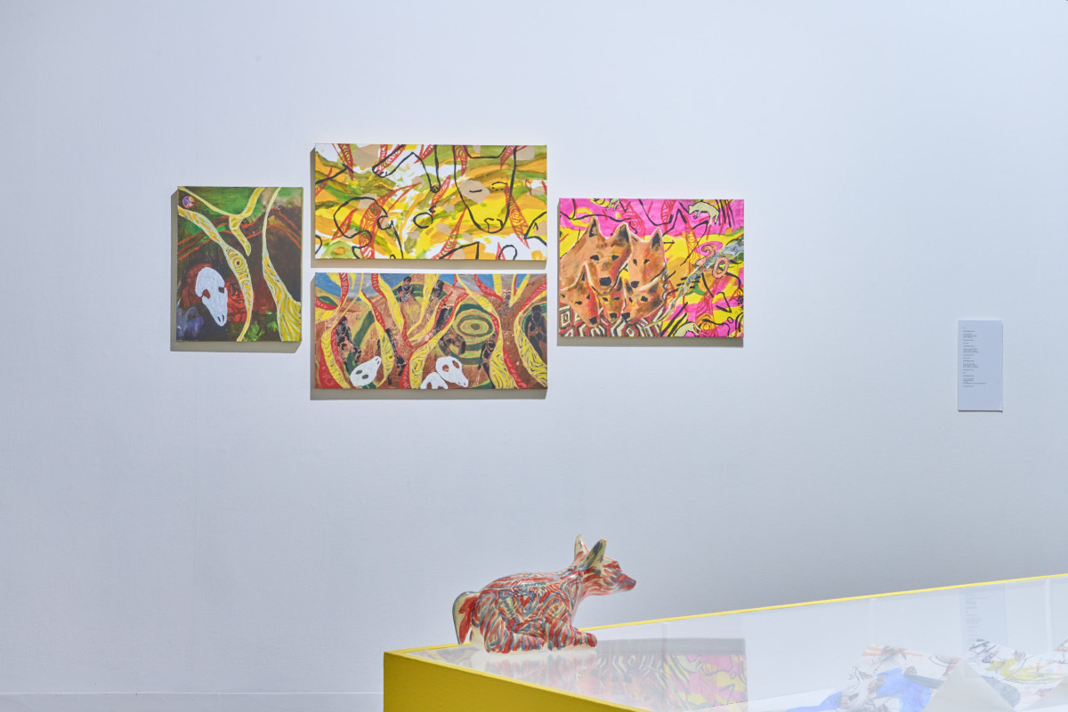 Four colourful artworks handing together on a white wall. A red ceramic dingo sits on a vitrine in the foreground.