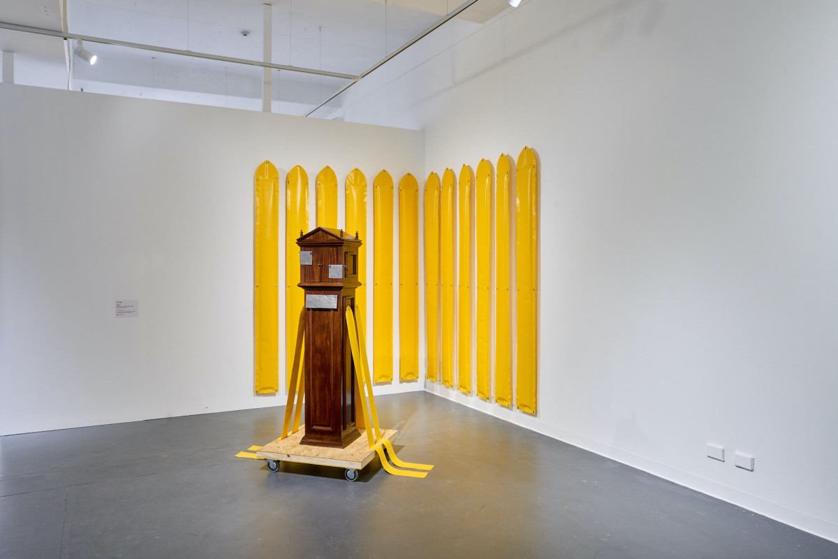 A wooden decorative reliquary sits on a dolly in the gallery. Many bright yellow radiation shielding panels hang on the white wall.