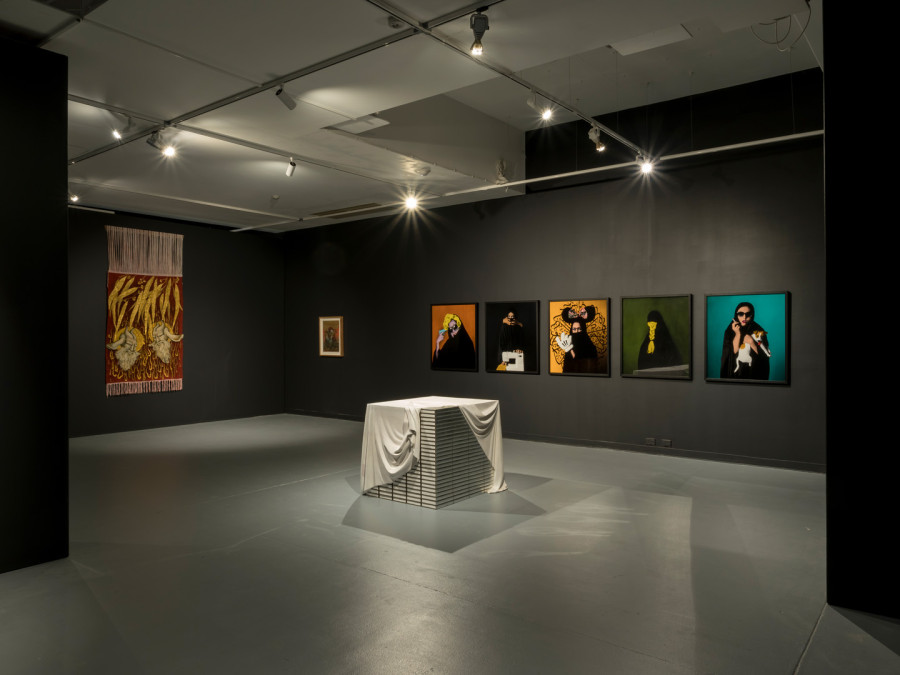A wide shot showing the inside of the exhibition - eight colourful artworks are shown, including a rich red and gold tapestery, a series of five pop-art inspired photographs and a wooden sculpture resembling a stack of silver books with a white sheet over them